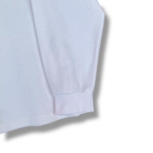 May include: Close-up of a white long-sleeve shirt. The cuff of the sleeve is ribbed and the fabric appears to be a soft knit. The shirt is plain and has no visible graphics or patterns.