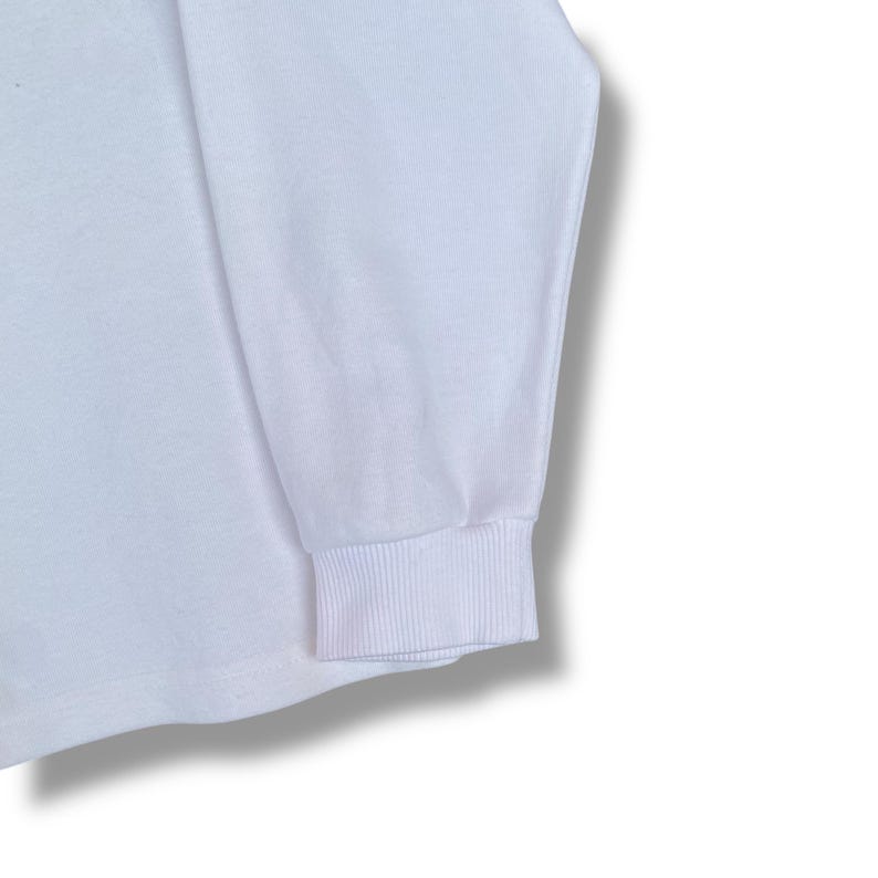 May include: Close-up of a white long-sleeve shirt. The cuff of the sleeve is ribbed and the fabric appears to be a soft knit. The shirt is plain and has no visible graphics or patterns.