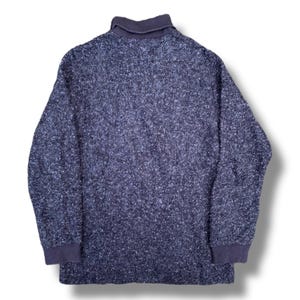 K&ouml;nnte beinhalten: Ein marineblauer Langarmpullover mit einem strukturierten, gesprenkelten Muster. Der Pullover hat einen dunkelblauen Kragen und Manschetten. Der Stoff scheint ein dickes Strickgewebe zu sein, das f&uuml;r k&uuml;hleres Wetter geeignet ist.