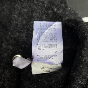 K&ouml;nnte beinhalten: Nahaufnahme eines schwarzen Kleidungsst&uuml;cketiketts mit japanischem Text und den Worten "MADE IN CHINA" in Wei&szlig;. Das Etikett ist an einem dunklen Stoff befestigt, m&ouml;glicherweise ein Pullover oder ein Mantel. Das Etikett enth&auml;lt Pflegehinweise.