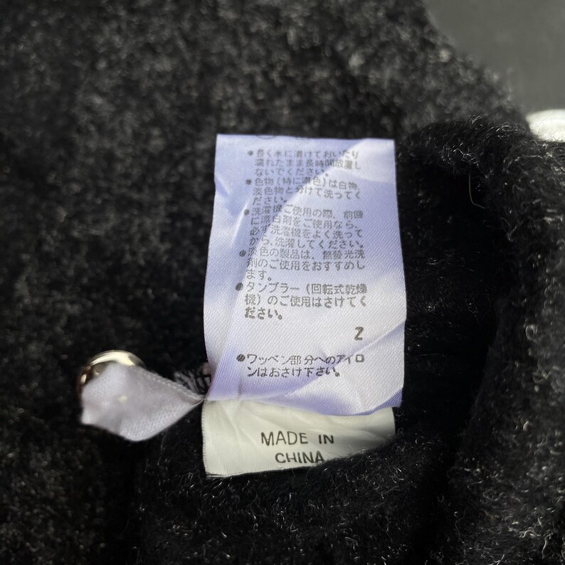 K&ouml;nnte beinhalten: Nahaufnahme eines schwarzen Kleidungsst&uuml;cketiketts mit japanischem Text und den Worten "MADE IN CHINA" in Wei&szlig;. Das Etikett ist an einem dunklen Stoff befestigt, m&ouml;glicherweise ein Pullover oder ein Mantel. Das Etikett enth&auml;lt Pflegehinweise.
