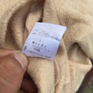 May include: A beige sweatshirt with a white tag that reads "ナイガイ" and includes the company's phone number, address, and size information. The tag also states that the sweatshirt is made of 80% acrylic and 20% polyester.