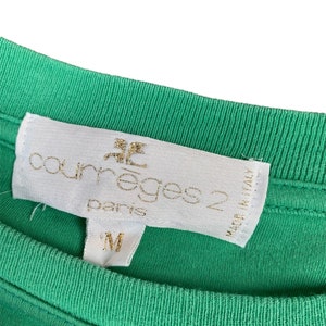 Puede incluir: Una etiqueta blanca con letras doradas que dice "Courreges 2 Paris" y "Made in Italy". La etiqueta est&aacute; unida a una camisa verde.