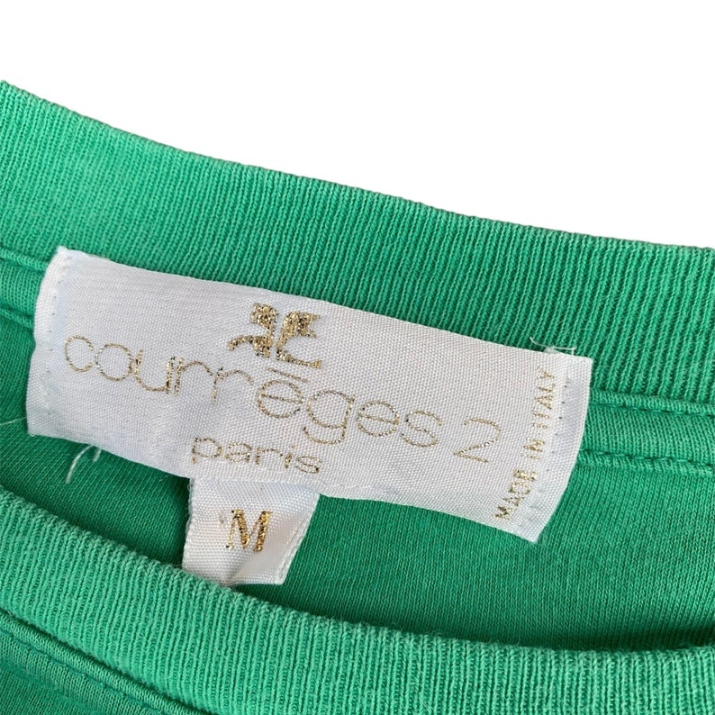 Puede incluir: Una etiqueta blanca con letras doradas que dice "Courreges 2 Paris" y "Made in Italy". La etiqueta est&aacute; unida a una camisa verde.