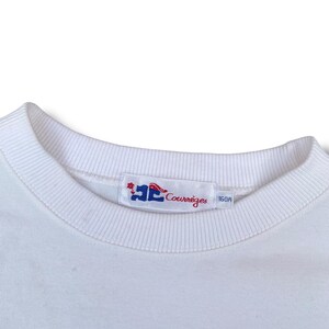 May include: Close-up of a white crew neck sweatshirt. The collar is ribbed. A rectangular tag with the brand name "Courr&egrave;ges" in red script and a logo is sewn on the collar. The tag also has the number "160A".