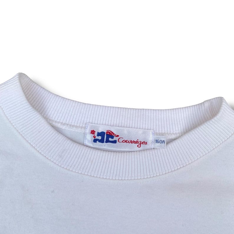 May include: Close-up of a white crew neck sweatshirt. The collar is ribbed. A rectangular tag with the brand name "Courr&egrave;ges" in red script and a logo is sewn on the collar. The tag also has the number "160A".