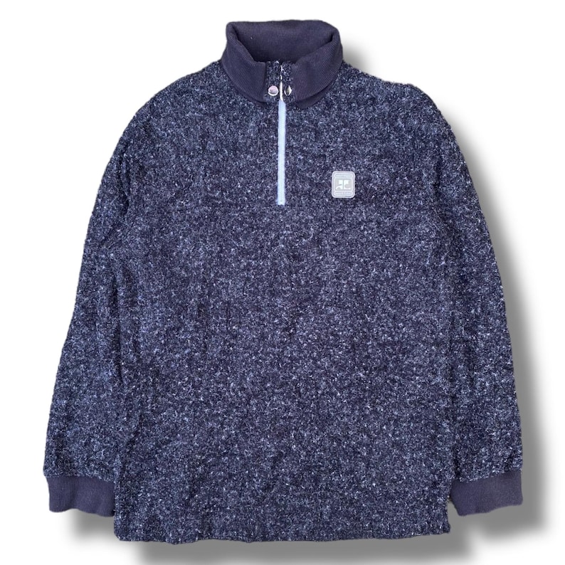 K&ouml;nnte beinhalten: Ein marineblauer Fleece-Pullover mit hohem Kragen und Viertelrei&szlig;verschluss. Der strukturierte Stoff hat wei&szlig;e Flecken. Ein kleines quadratisches Emblem befindet sich auf der Brust. Die Manschetten und der Kragen sind in einem dunkleren Marineblau gehalten.