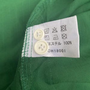May include: Close-up of a green fabric with two white buttons and a tag. The tag has care symbols and text that reads "エステル 100%" and "CH18001". The fabric has a subtle texture.