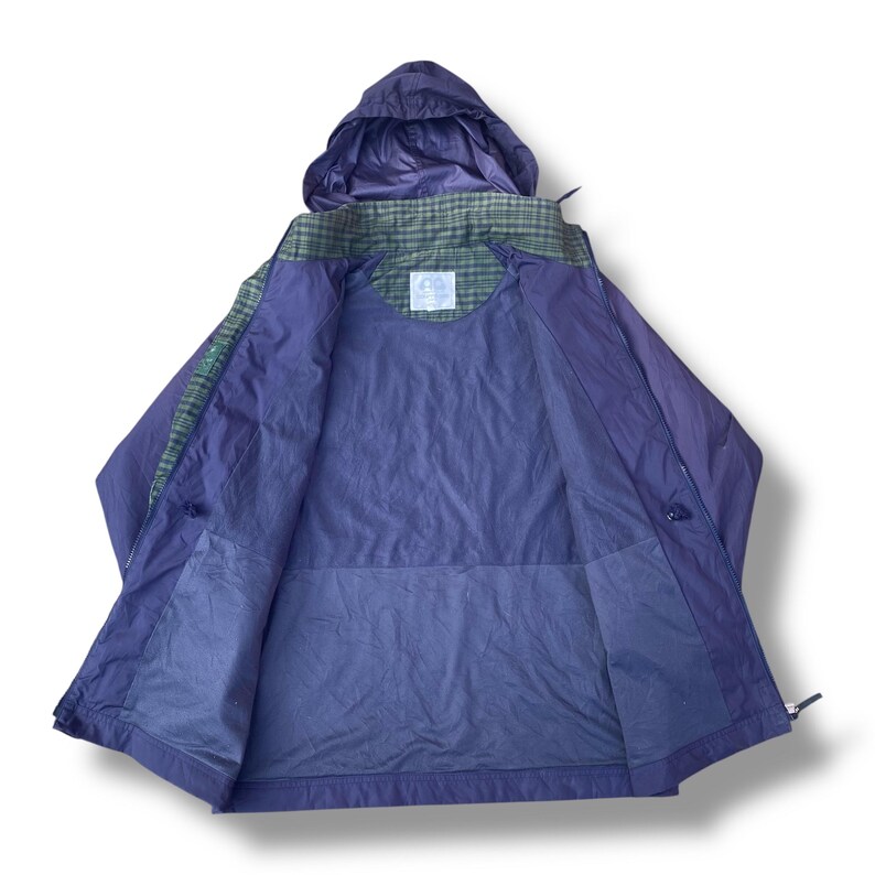May include: A purple and green hooded jacket, open to display the interior lining. The jacket features a plaid pattern on the shoulders and hood, with a solid purple body. The jacket has a zipper closure and a small logo patch.