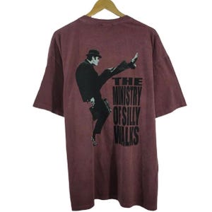 May include: A maroon short-sleeve t-shirt featuring a black graphic of a person in a suit and bowler hat performing a high kick. The text "THE MINISTRY OF SILLY WALKS" is printed in bold black capital letters next to the figure on the back of the shirt.