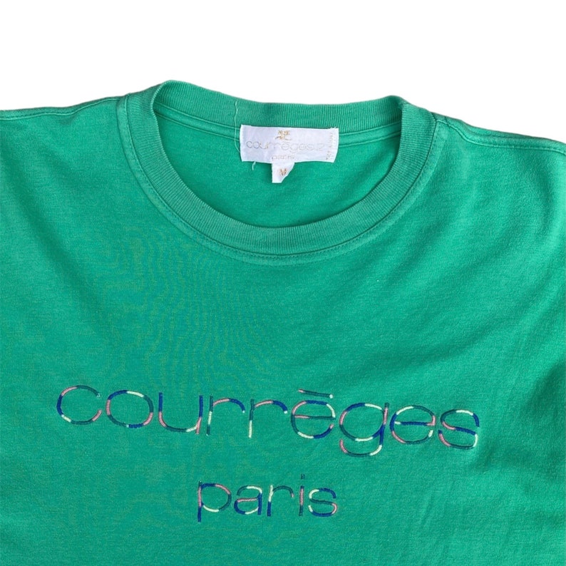 Puede incluir: Camiseta verde con una etiqueta blanca que dice "Courr&egrave;ges Paris" y un logotipo bordado que dice "Courr&egrave;ges Paris" en azul, rosa y blanco.