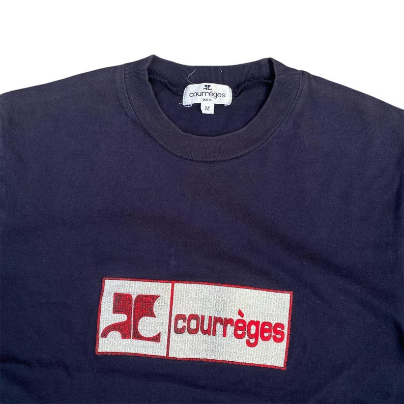 K&ouml;nnte beinhalten: Ein dunkelblaues T-Shirt mit Rundhalsausschnitt und einem rechteckigen Aufdruck auf der Brust. Der Aufdruck zeigt ein rotes Logo und den Schriftzug "courr&egrave;ges" in roten Buchstaben. Ein wei&szlig;es Innenetikett am Kragen zeigt "courr&egrave;ges" und "M".
