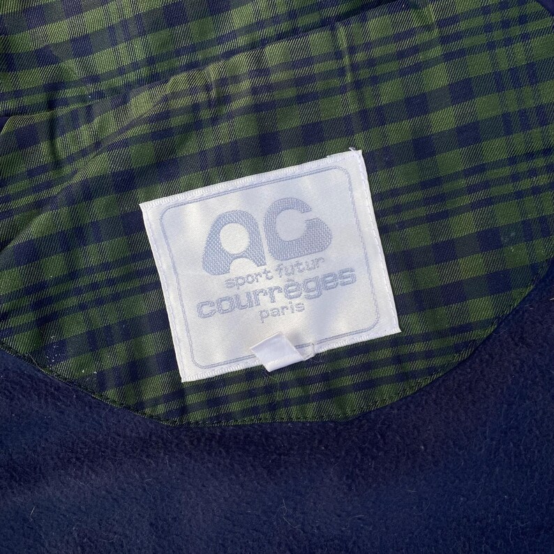 May include: Close-up of a navy blue garment with a green and navy plaid lining. A white label with the text "AC sport futur courr&egrave;ges paris" is sewn into the garment.