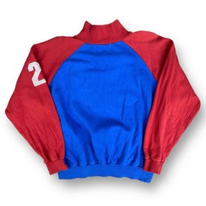 Peut inclure: Un sweat-shirt vintage avec des blocs de couleurs rouge, bleu et blanc. Les manches raglan sont rouges et le corps est bleu. Le col montant et les poignets sont rouges. Le chiffre blanc "2" est sur la manche gauche. Ce sweat-shirt de style r&eacute;tro est parfait pour un usage quotidien.