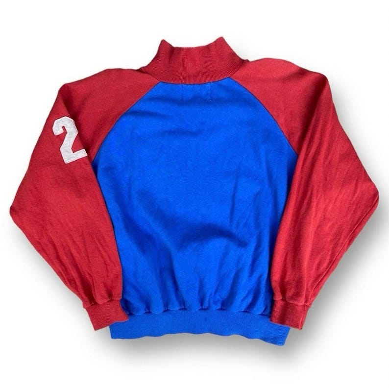 Peut inclure: Un sweat-shirt vintage avec des blocs de couleurs rouge, bleu et blanc. Les manches raglan sont rouges et le corps est bleu. Le col montant et les poignets sont rouges. Le chiffre blanc "2" est sur la manche gauche. Ce sweat-shirt de style r&eacute;tro est parfait pour un usage quotidien.