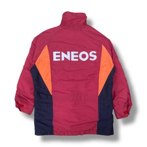 Può includere: Giacca rossa e arancione con la scritta "ENEOS" a lettere bianche sul retro. La giacca ha un collo alto e pannelli laterali blu scuro. Le maniche hanno chiusure in velcro rosse.