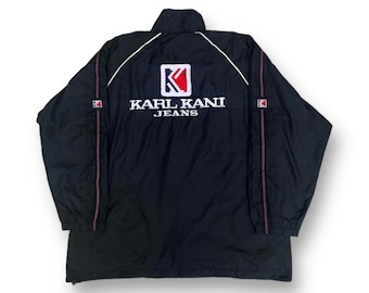 May include: Black jacket with white and red accents. The back features the Karl Kani Jeans logo in white, with a red and white square logo above the text. The jacket has a high collar and a zipper closure. The jacket has red stitching on the sleeves.