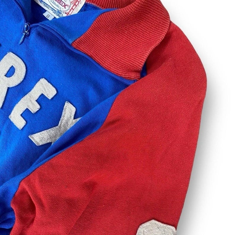 Peut inclure: Un sweat-shirt zipp&eacute; bleu et rouge avec le mot "REX" en lettres blanches. Le sweat-shirt a un col et des manches rouges et un corps bleu. La fermeture &eacute;clair est argent&eacute;e. L'&eacute;tiquette indique "REX Classics".