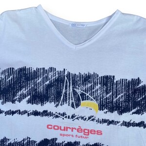 K&ouml;nnte beinhalten: Wei&szlig;es T-Shirt mit einem blauen und wei&szlig;en Grafikdruck einer Bergkette und einer gelben Sonne. Der Text "courr&egrave;ges sport futur" ist in Pink unter dem Grafikdruck gedruckt.