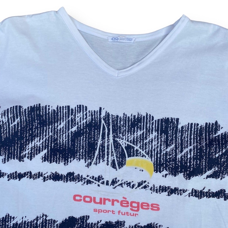 K&ouml;nnte beinhalten: Wei&szlig;es T-Shirt mit einem blauen und wei&szlig;en Grafikdruck einer Bergkette und einer gelben Sonne. Der Text "courr&egrave;ges sport futur" ist in Pink unter dem Grafikdruck gedruckt.