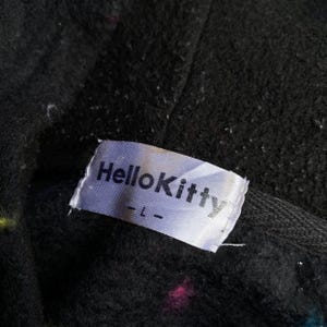 Puede incluir: Primer plano de una tela negra con una etiqueta blanca que dice "Hello Kitty" en texto negro. La etiqueta tambi&eacute;n tiene la talla "-L-" impresa. La tela tiene motas de color.