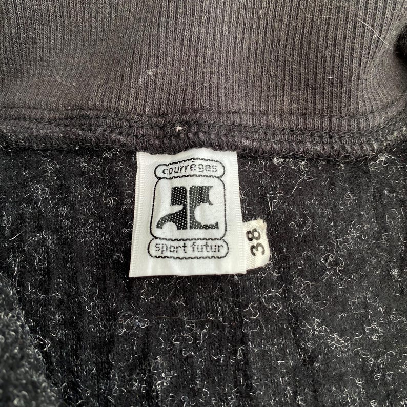 K&ouml;nnte beinhalten: Nahaufnahme eines schwarzen Kleidungsst&uuml;cks mit gerippter Struktur. Ein wei&szlig;es Etikett zeigt den Markennamen "courr&egrave;ges" &uuml;ber einem Logo und die Worte "sport futur". Die Nummer "38" ist auf einem separaten Etikett aufgedruckt.