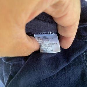 K&ouml;nnte beinhalten: Ein dunkelblaues Kleidungsst&uuml;ck mit einem wei&szlig;en, rechteckigen Pflegeetikett, das den schwarzen Text 100% COTTON, MACHINE WASH COLD, DELICATE CYCLE, LINE DRY, COOL IRON IF NECESSARY, WASH SEPARATELY, DO NOT BLEACH und DO NOT DRY CLEAN enth&auml;lt.