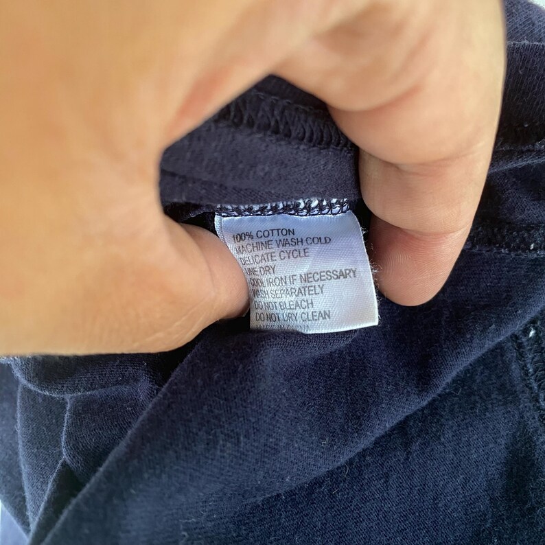 K&ouml;nnte beinhalten: Ein dunkelblaues Kleidungsst&uuml;ck mit einem wei&szlig;en, rechteckigen Pflegeetikett, das den schwarzen Text 100% COTTON, MACHINE WASH COLD, DELICATE CYCLE, LINE DRY, COOL IRON IF NECESSARY, WASH SEPARATELY, DO NOT BLEACH und DO NOT DRY CLEAN enth&auml;lt.