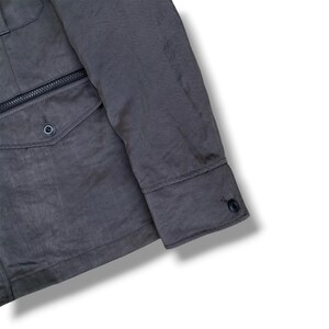 K&ouml;nnte beinhalten: Eine dunkelgraue Jacke mit einem langen &Auml;rmel mit gekn&ouml;pfter Manschette, einer Vordertasche mit Klappe und einem einzelnen Knopfverschluss sowie einer horizontalen Rei&szlig;verschlusstasche direkt &uuml;ber der Klappentasche auf der Vorderseite.