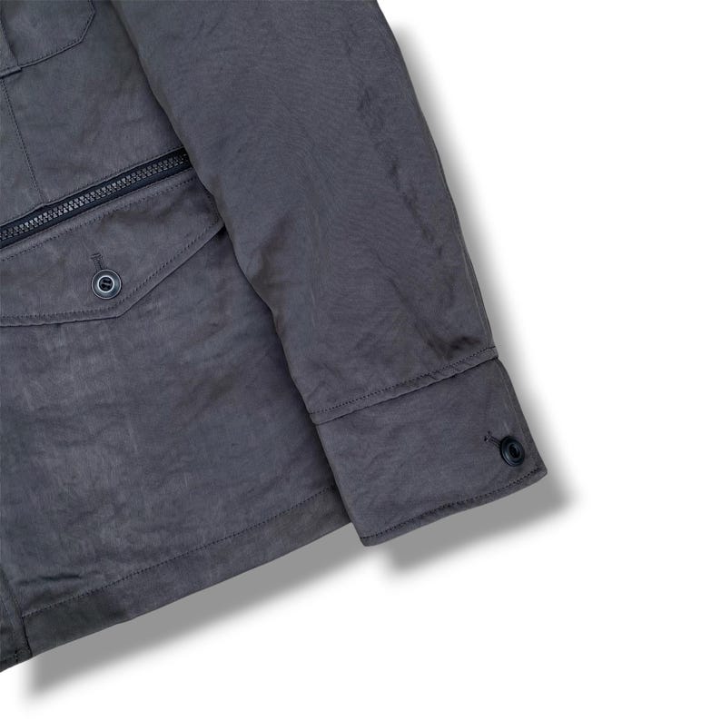 K&ouml;nnte beinhalten: Eine dunkelgraue Jacke mit einem langen &Auml;rmel mit gekn&ouml;pfter Manschette, einer Vordertasche mit Klappe und einem einzelnen Knopfverschluss sowie einer horizontalen Rei&szlig;verschlusstasche direkt &uuml;ber der Klappentasche auf der Vorderseite.