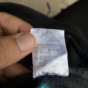 Puede incluir: Primer plano de una etiqueta blanca con texto japon&eacute;s e ingl&eacute;s, incluyendo "FOR SALE IN JAPAN ONLY". La etiqueta est&aacute; adherida a una tela negra. La etiqueta tiene forma rectangular.