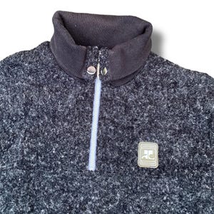 K&ouml;nnte beinhalten: Ein dunkelgrauer Fleece-Pullover mit hohem, geripptem Kragen und einem Viertel-Rei&szlig;verschluss. Der Rei&szlig;verschluss ist hellblau. Ein kleines Patch mit dem Text "Sport Futur" befindet sich auf der Brust.