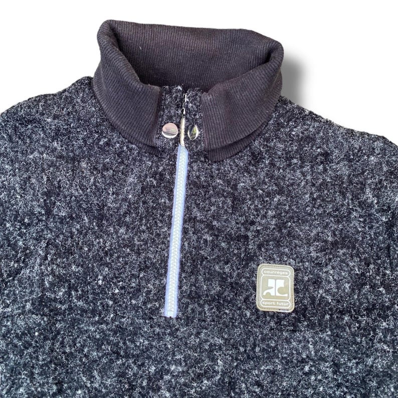 K&ouml;nnte beinhalten: Ein dunkelgrauer Fleece-Pullover mit hohem, geripptem Kragen und einem Viertel-Rei&szlig;verschluss. Der Rei&szlig;verschluss ist hellblau. Ein kleines Patch mit dem Text "Sport Futur" befindet sich auf der Brust.