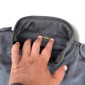 K&ouml;nnte beinhalten: Eine dunkelolivgr&uuml;ne Jacke mit hohem Stehkragen und einem kleinen Innenetikett mit der Aufschrift A.A.R. Das Kleidungsst&uuml;ck besteht aus einem strapazierf&auml;higen, dicht gewebten Stoff mit sichtbaren N&auml;hten entlang des Kragens und der Schulterklappen.