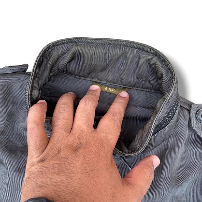 K&ouml;nnte beinhalten: Eine dunkelolivgr&uuml;ne Jacke mit hohem Stehkragen und einem kleinen Innenetikett mit der Aufschrift A.A.R. Das Kleidungsst&uuml;ck besteht aus einem strapazierf&auml;higen, dicht gewebten Stoff mit sichtbaren N&auml;hten entlang des Kragens und der Schulterklappen.