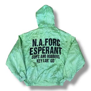 Puede incluir: Chaqueta con capucha verde claro con texto negro en la espalda. El texto dice "N.A. FORC ESPERANT COPS AND ROBBERS KEY LAR' GO". La chaqueta tiene un cordón en la parte inferior y una capucha.
