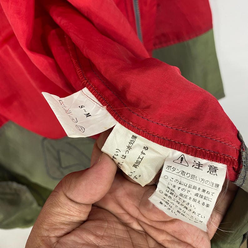Op de afbeelding: Een rood en groen jack met een ritssluiting. Het jack heeft een label met de tekst "S~M" en een waarschuwingslabel in het Japans dat "注意 ポタン取り扱い方の注意" zegt, wat vertaald wordt als "Let op: Instructies voor het omgaan met knoppen".