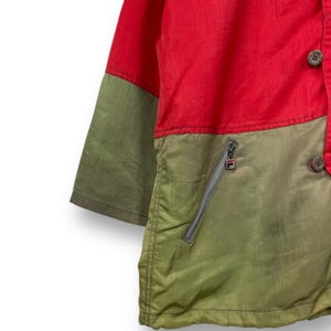 Op de afbeelding: Een rood, groen en olijfgroen jack met een ritszak en een knoopsluiting. Het jack heeft een contrasterend kleurendesign.