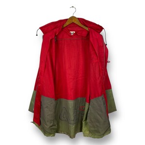 Op de afbeelding: Een rood en groen jack met een ritssluiting en een capuchon. Het jack heeft een groot geborduurd logo op de rug.