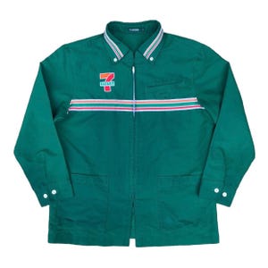 May include: A green button-up jacket with a white collar and a zipper closure. The jacket features the 7-Eleven logo on the chest and a horizontal stripe design in red, orange, and white. Two front pockets and white buttons complete the design.
