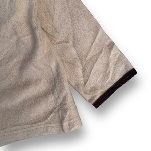 May include: A cream-colored fleece jacket with a brown trim around the edges. The jacket has a textured surface and a relaxed fit.