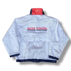 May include: White nylon jacket featuring the text "AICHI TOYOTA" in red and navy blue. The jacket has a red collar with the Toyota logo and navy blue trim at the cuffs and hem. A vintage-style jacket.