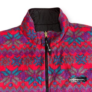 May include: A red fleece jacket with a black zipper and a colorful geometric pattern. The jacket has a black lining and a small patch with the text "Storm" on the bottom right side.