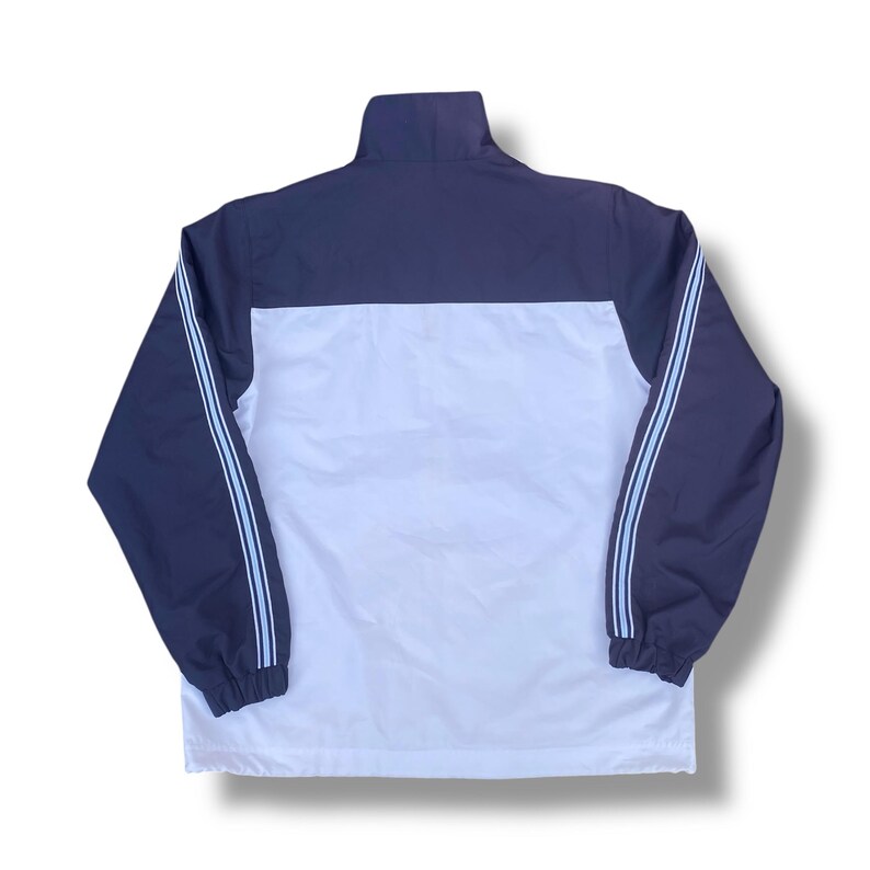 May include: A navy blue and white track jacket with a high collar. The jacket features light blue stripes down the sleeves and a white panel across the chest. The jacket has a full zipper and elastic cuffs.