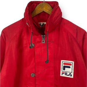 Op de afbeelding: Rode Fila jas met ritssluiting, capuchon en een zwarte Fila logo patch aan de rechterkant van de borst.
