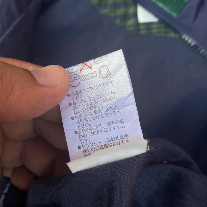 May include: A close-up of a white clothing tag with Japanese text and laundry symbols. The tag is held by a hand and is attached to a navy blue garment. The text provides washing instructions.
