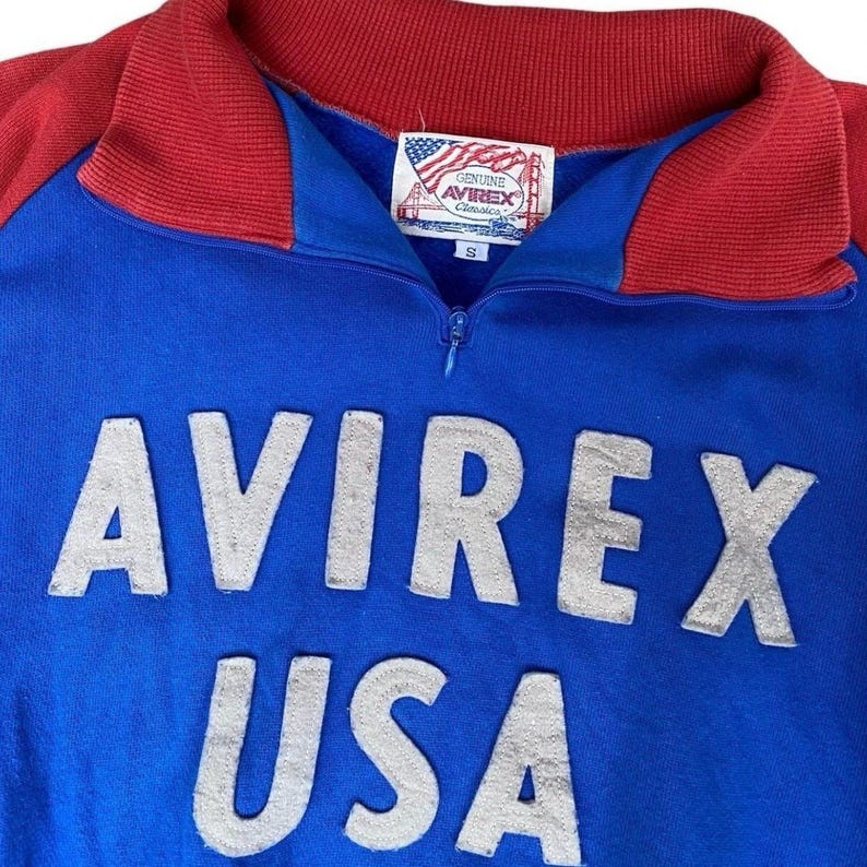 Peut inclure: Un sweat-shirt bleu et rouge &agrave; fermeture &eacute;clair avec le texte "AVIREX USA" en grandes lettres argent&eacute;es. Le sweat-shirt a un col et des &eacute;paules rouges. Une petite &eacute;tiquette avec le drapeau am&eacute;ricain et les mots "GENUINE AVIREX Classics" est cousue sur la poitrine.