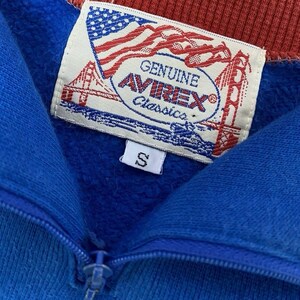 Peut inclure: Gros plan d'un sweat-shirt bleu et rouge &agrave; fermeture &eacute;clair. Le sweat-shirt porte une &eacute;tiquette avec le texte "GENUINE AVIREX Classics" et un motif de drapeau am&eacute;ricain. Une petite &eacute;tiquette blanche avec la lettre "S" est visible.