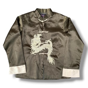May include: Olive green satin jacket with a mandarin collar and frog closures. The jacket features a large embroidered dragon design in cream-colored thread on the front. The cuffs are trimmed with a cream-colored satin.