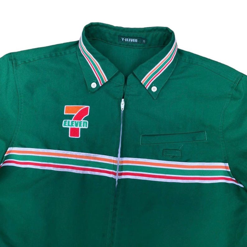 May include: Green button-down shirt with a white, red, and orange stripe across the chest and collar. The shirt features the 7-Eleven logo on the left chest and the text "7-ELEVEN" on the collar.